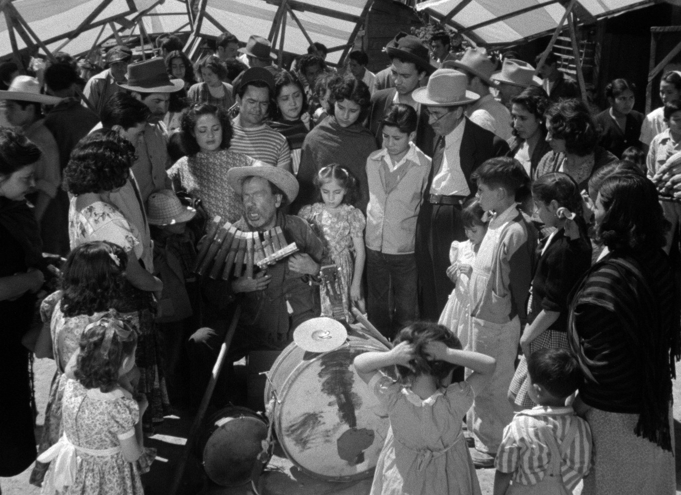 Fotograma de Los olvidados: un grupo de vecinos y niños rodea a un músico callejero